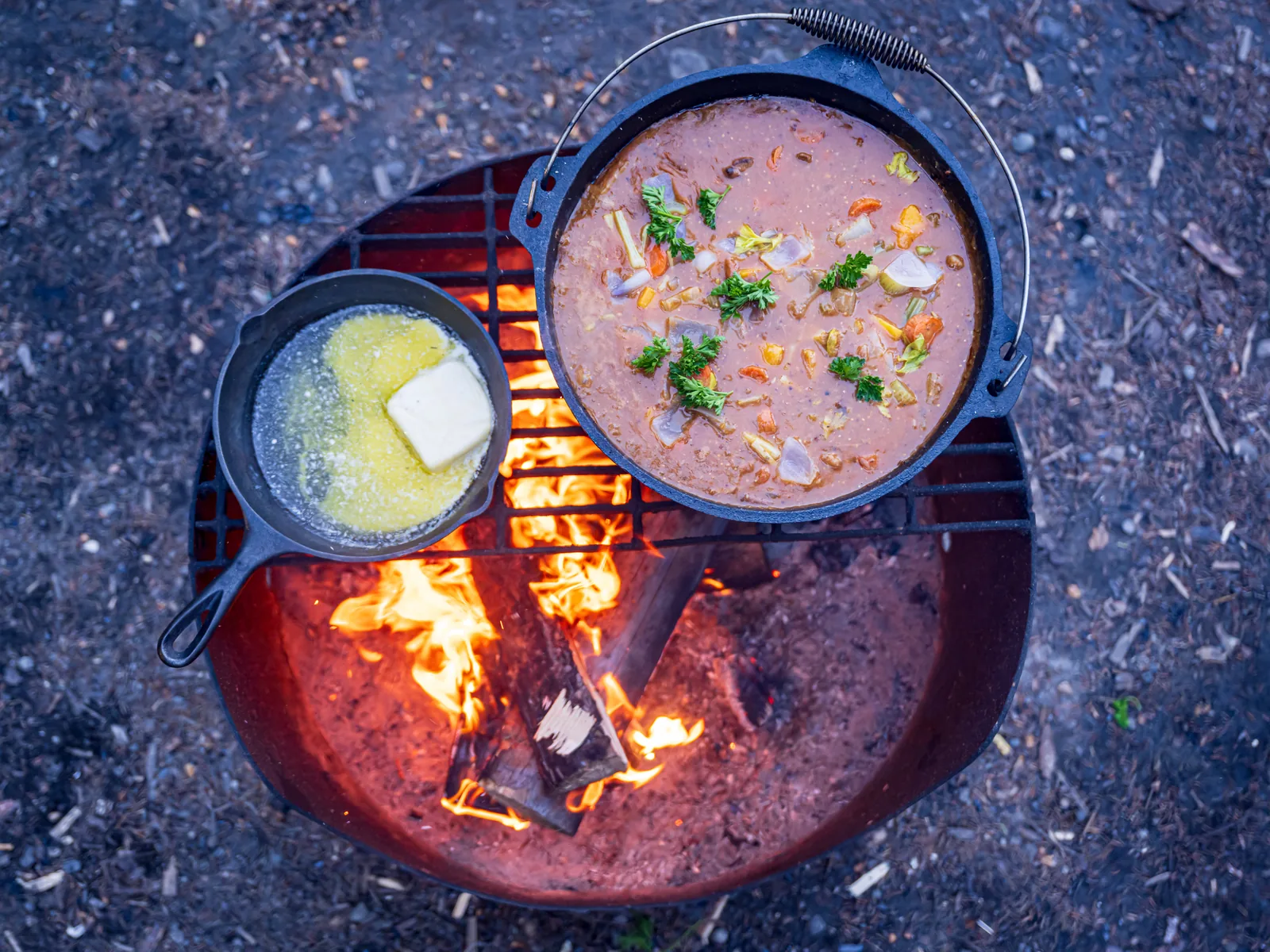 Overhead view of a Dutch oven of stew and a skillet of melting butter over an open fire pit