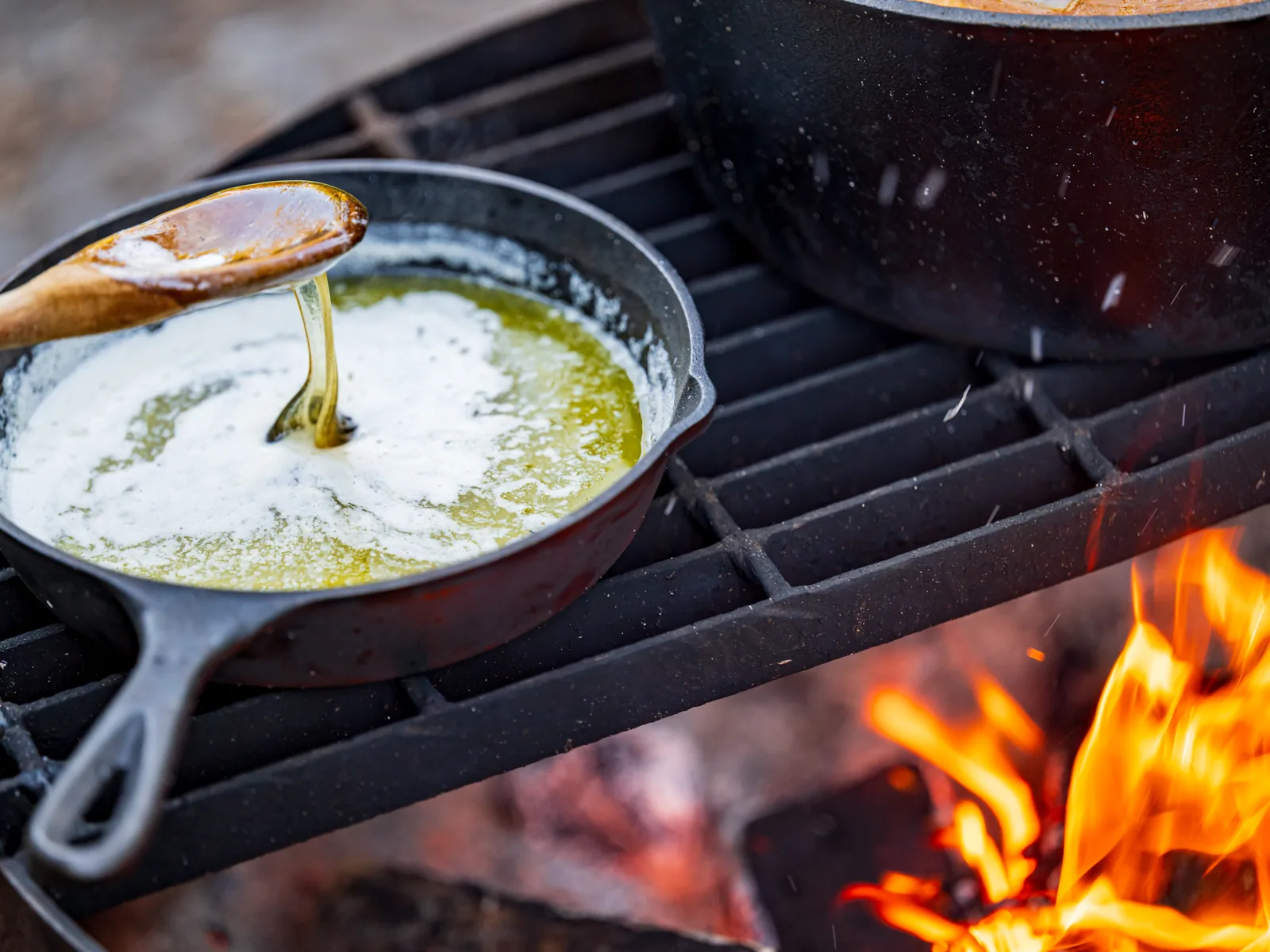 A cast-iron skillet of melted butter over live flames on a fire grate, stirred with a wooden spoon