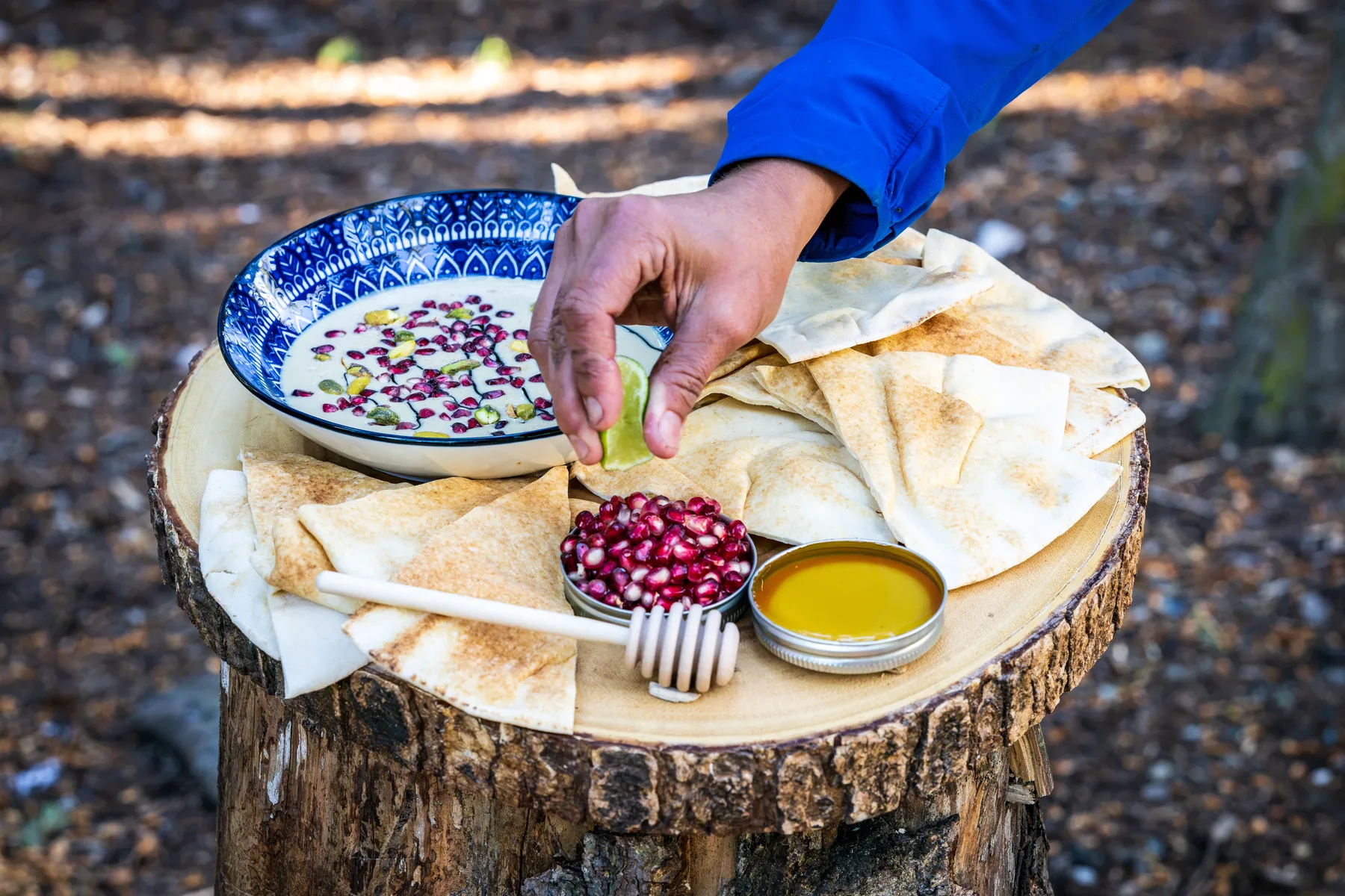 A hand adding a squeeze of lime to a bowl of dip beside flatbread, pomegranate seeds and honey on a tree stump in a sunlit forest