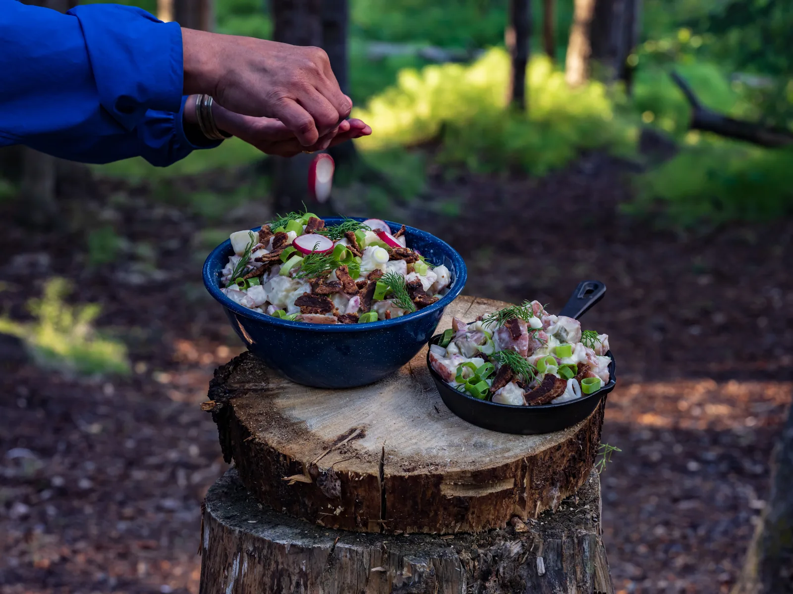 A hand adding a sliced radish to a bowl of salad on a tree stump in the forest